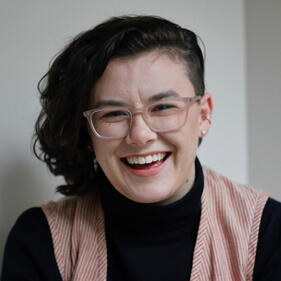 a headshot of a white woman with chin length brown curly hair. she is wearing light pink glasses, a black turtleneck, and a vest